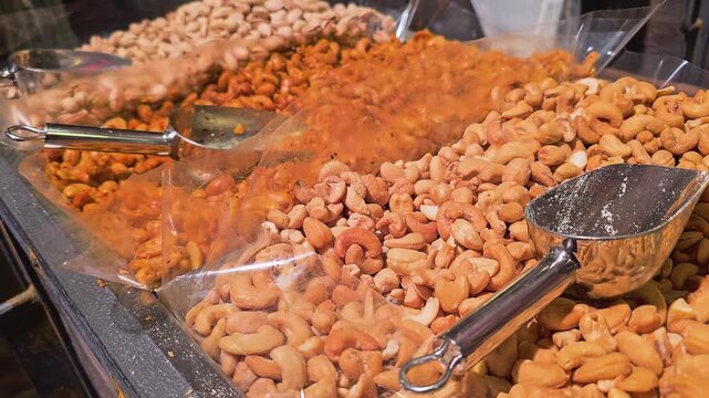 Close view across rows of roasted nuts and snack mixes in a market case, with reflective metal scoops, clear bins, warm lights, and textured bulk food retail presentation.