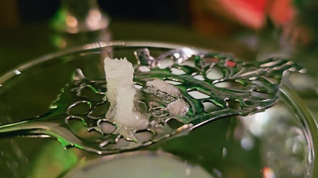 Macro close-up of sugar cube dissolving on ornate absinthe spoon, with bubbles, milky louche below, green bar reflections, shallow focus, and classic cocktail ritual atmosphere.
