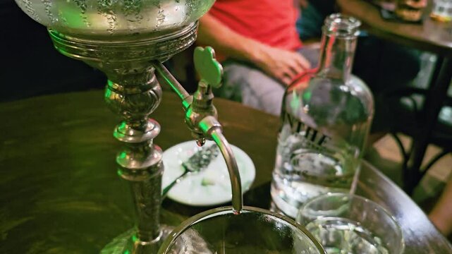Close-up of absinthe louche dripping from a vintage fountain spout into a glass, with liquor bottle, water glass, slotted spoon, round bar table, and moody green lounge lighting.