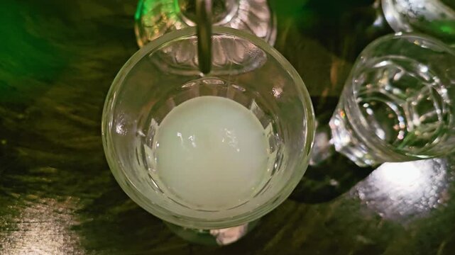 Close-up of absinthe turning milky as water drops into a glass from an ornate fountain, with green bar lighting, glossy tabletop, and classic cocktail service atmosphere.