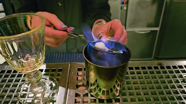 Absinthe ritual close-up with blue flame curling around a sugar cube on a perforated spoon, metal cup, goblet glass, bartender tools, stainless counter, and moody bar background.