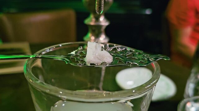 Close-up cocktail service showing sugar cube on absinthe spoon over a milky drink, with fountain spout, water drip, glass reflections, and moody bar table scene at night.