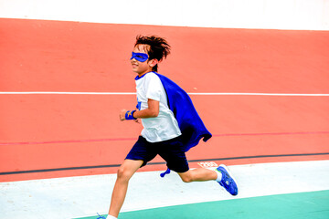 Fototapeta premium Child in superhero costume running on a track, wearing a blue cape.