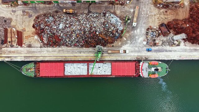 A cargo ship is seen at a port as it gets loaded with materials. Nearby, there are large piles of scrap metal and construction equipment. Several trucks are visible in the area.
