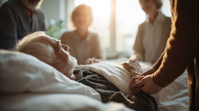 An elderly man lies in a hospital bed, surrounded by supportive family. Warm sunlight and gentle hands convey compassion, love, and end-of-life care.