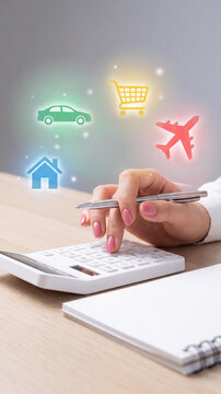 Close-up of a woman hand using a white calculator on a desk next to a notebook, with glowing icons of a house, car, shopping cart, and plane symbolizing family budget planning.