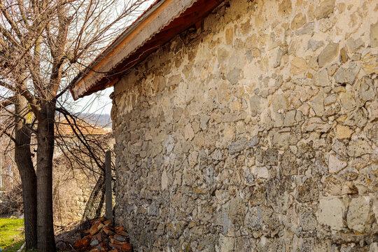 An old stone house wall with a red-tiled roof stands beside bare trees, its rugged rock texture weathered by time in a rural setting