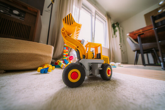 Yellow toy front loader construction truck photographed at low angle on a home carpet, surrounded by colorful building blocks - a scene of imaginative childhood play and family life