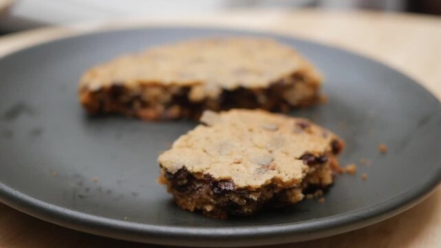 A person picks up and breaks a freshly baked homemade chocolate chip cookie on a dark plate, showing its soft and chewy texture with delicious chocolate chunks ready to be enjoyed as a sweet treat