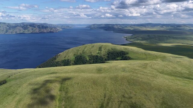 A breathtaking aerial drone flight over a peaceful mountain bay framed by rolling green hills and expansive freshwater. Towering clouds cast dynamic light across the landscape, creating a dramatic sum