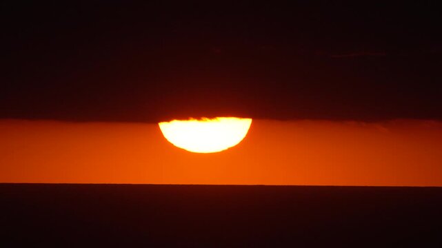 Intense orange sun rests below a dark cloud bank over the ocean horizon at dusk. Static fixed shot.