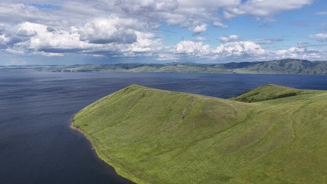 A breathtaking aerial drone flight over a peaceful mountain bay framed by rolling green hills and expansive freshwater. Towering clouds cast dynamic light across the landscape, creating a dramatic sum