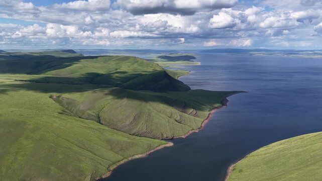 A breathtaking aerial drone flight over a peaceful mountain bay framed by rolling green hills and expansive freshwater. Towering clouds cast dynamic light across the landscape, creating a dramatic sum