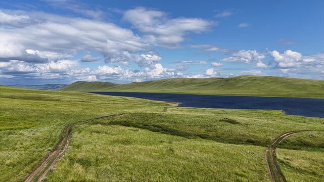 Cinematic drone footage of a tranquil mountain lake stretching between rolling green hills beneath dramatic cumulus clouds. Scenic aerial landscape featuring pristine nature, deep blue water, panorami