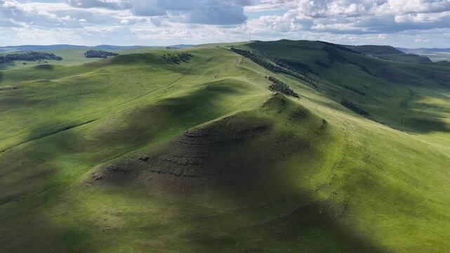 Cinematic drone footage soaring above rolling green hills under a dramatic cloudy sky. Soft sunlight and moving cloud shadows create striking patterns across the natural landscape, revealing the beaut