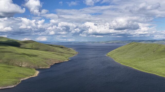 Cinematic drone footage of a tranquil mountain lake stretching between rolling green hills beneath dramatic cumulus clouds. Scenic aerial landscape featuring pristine nature, deep blue water, panorami