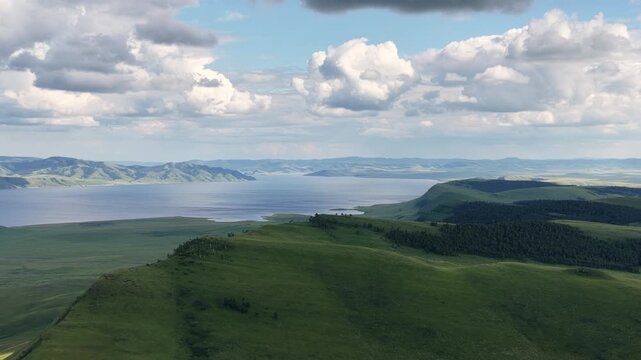 Spectacular drone footage capturing a vast mountain lake bordered by rolling green highlands and forested slopes beneath towering clouds. The changing light creates a peaceful yet dramatic atmosphere,