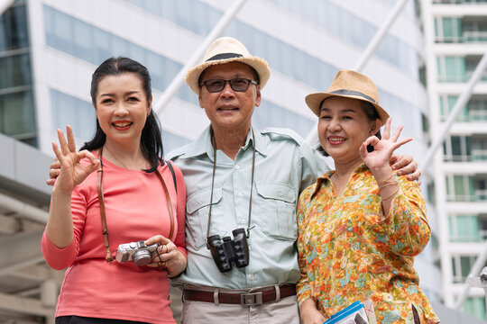 Happy Asian family enjoying a city vacation. Senior parents and adult daughter smiling, showing OK hand signs with a camera and binoculars in a modern urban setting.