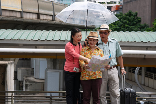 Happy Asian family traveling in the city during a cloudy day. Adult daughter holding a clear umbrella while helping her senior parents read a map to find directions on an urban skywalk.