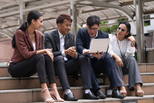 Group of diverse Asian business professionals sitting on outdoor city stairs looking at a laptop. Happy corporate team collaborating, discussing a project, and working outside the office.