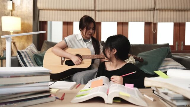 Two Asian teenage tutorials, girl concentrates on reading books for exam, learning at home, her friend relaxes by playing guitar music, self-study with textbooks, homeschooling together lifestyle.