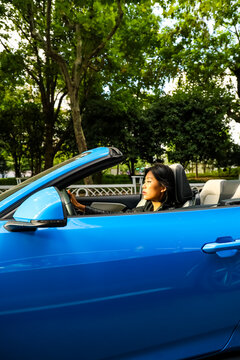 An Asian woman drives a blue convertible sports car down the street.