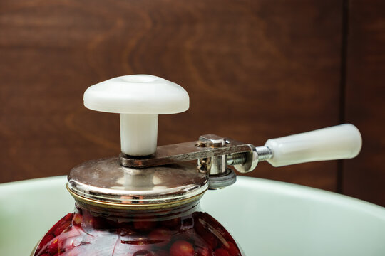 A Can Seamer. Canning cherry compote using a manual seaming machine to seal a large glass jar with metal lid. Home food preservation, traditional kitchen, winter storage preparations. Can in a basin.