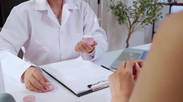 Asian woman doctor explaining and consult with young female patient in medical clinic applying cosmetic moisture cream product on her hand, computer screen showing ai facial skin analysis data.