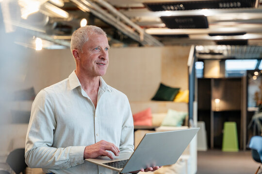 Smiling businessman holding laptop standing in modern office