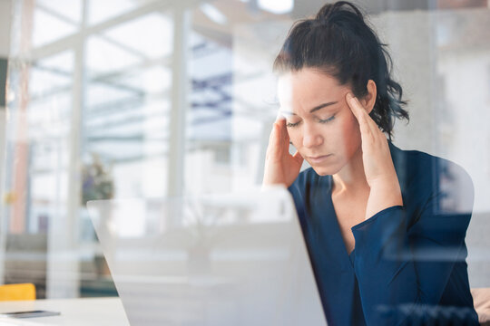 Businesswoman with eyes closed sitting in front of laptop seen through glass