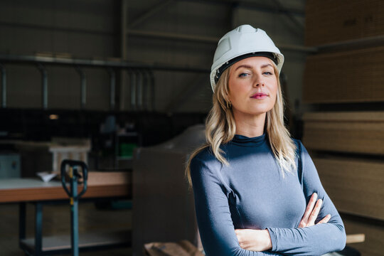 Confident businesswoman with arms crossed wearing hardhat in carpentry factory