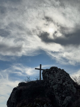 Wooden cross on rocky hilltop under cloudy sky with sunlight breaking through.