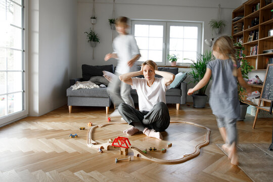 Stressed out mother sitting in the middle of toys, while children are running around her