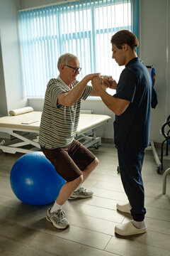 Male physiotherapist helps senior patient with squat exercise indoors