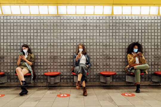 Friends with protective face mask using mobile phone while sitting at social distance in subway