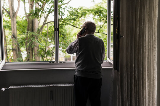 Back view of senior man on the phone standing at open window