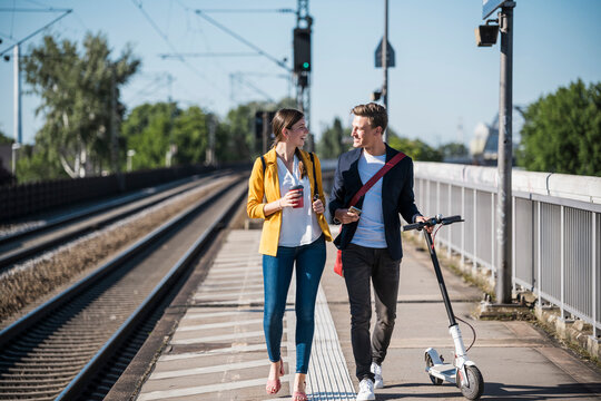Young couple walks on station platform with scooter and coffee
