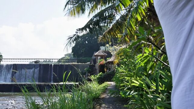 Man running while carrying the Indonesian flag near Tukad Unda Waterfall in Klungkung, Bali. Patriotic outdoor scene with tropical scenery, cascading water, and lush greenery.