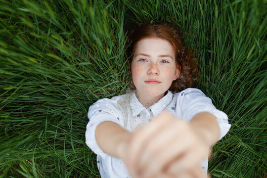 A girl with red hair reaches her hand toward the camera in the grass
