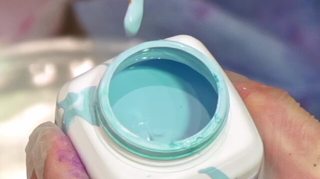 A gloved hand gently dips a golden-bristled brush into a pot of vivid aqua paint. The smooth, liquid color swirls as the brush is immersed, ready for artistic creation. A perfect moment.