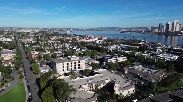High angle view highlights sunlit tennis complexes, lush greenery, resort architecture positioned directly along harbor edge, overlooking vibrant downtown San Diego skyline in Coronado, California