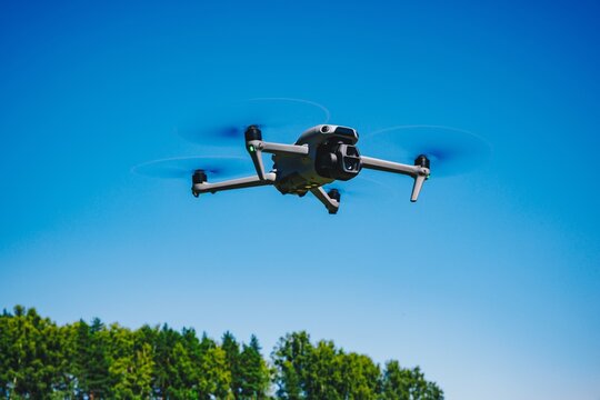 Agricultural or filming drone flying high in the blue sky above green forest treetops