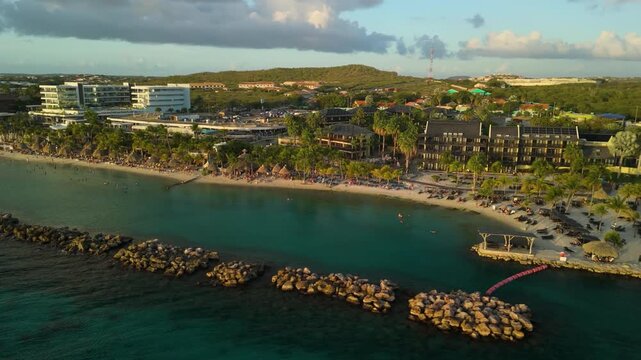 Aerial orbit reveal shot of Mambo Beach at sunset in Curacao with resorts, palm trees, breakwater rocks and golden light