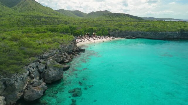 Aerial flight along rocky cliff coastline toward Kleine Knip Beach in Curacao revealing turquoise bay, green hills, thatched umbrellas, sunbathers, swimmers and snorkelers below