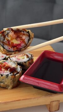 Golden fried sushi rolls are carefully dipped into rich soy sauce using wooden chopsticks above a red rectangular dipping dish