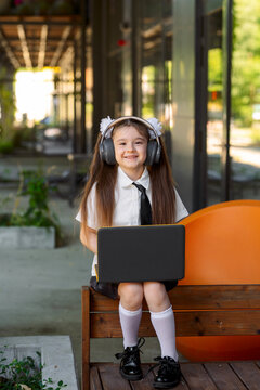Smiling schoolgirl wearing headphones using a laptop while sitting outdoors in a modern campus setting. Online learning, digital education, remote study and technology for children concept.