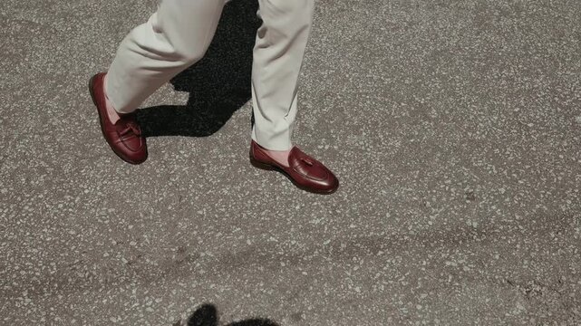 Legs and burgundy leather loafers cast long shadow on sunny cracked pavement outdoors