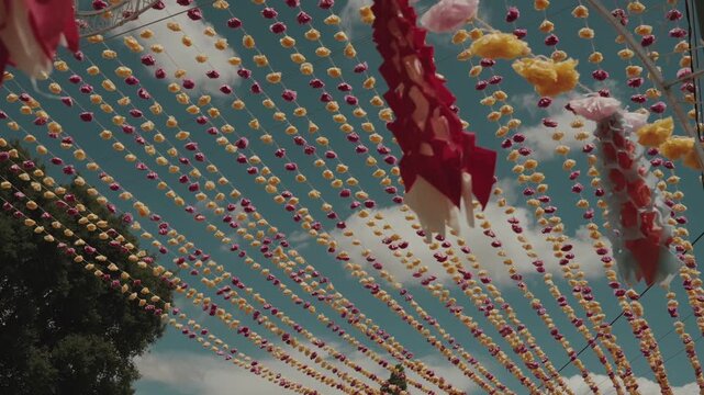 Dense rows of paper flower beads hang above tree canopy against blue sky at arraial festival in Portugal