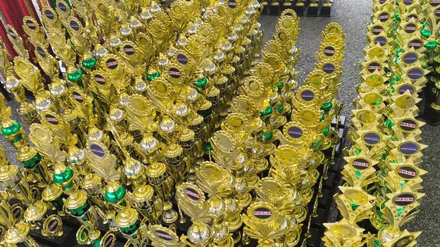 Many gold trophies are arranged neatly on the floor. The organizing committee sorted them by competition category. Their shining, elegant designs await the proud tournament winners.