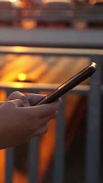 Young woman's hands holding and using a mobile phone, texting a message or browsing social media on a city bridge during a warm sunset, with out of focus car lights creating bokeh